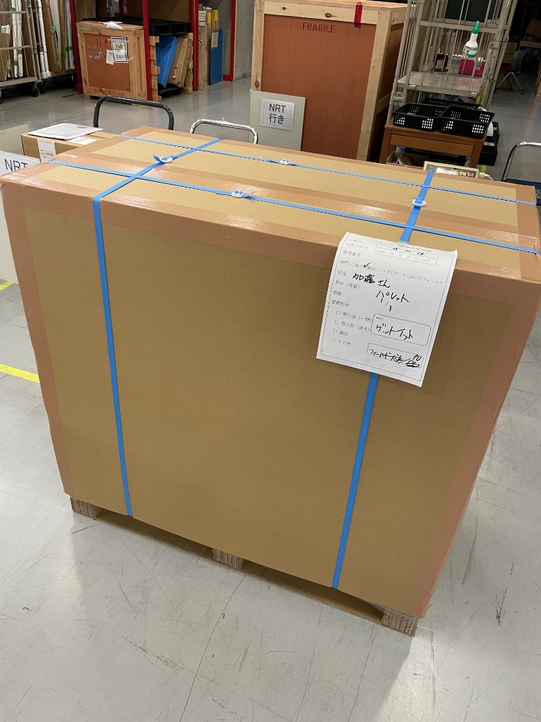 A consignment crated and strapped on a pallet in a Japanese cargo warehouse, beside a sign reading NRT.