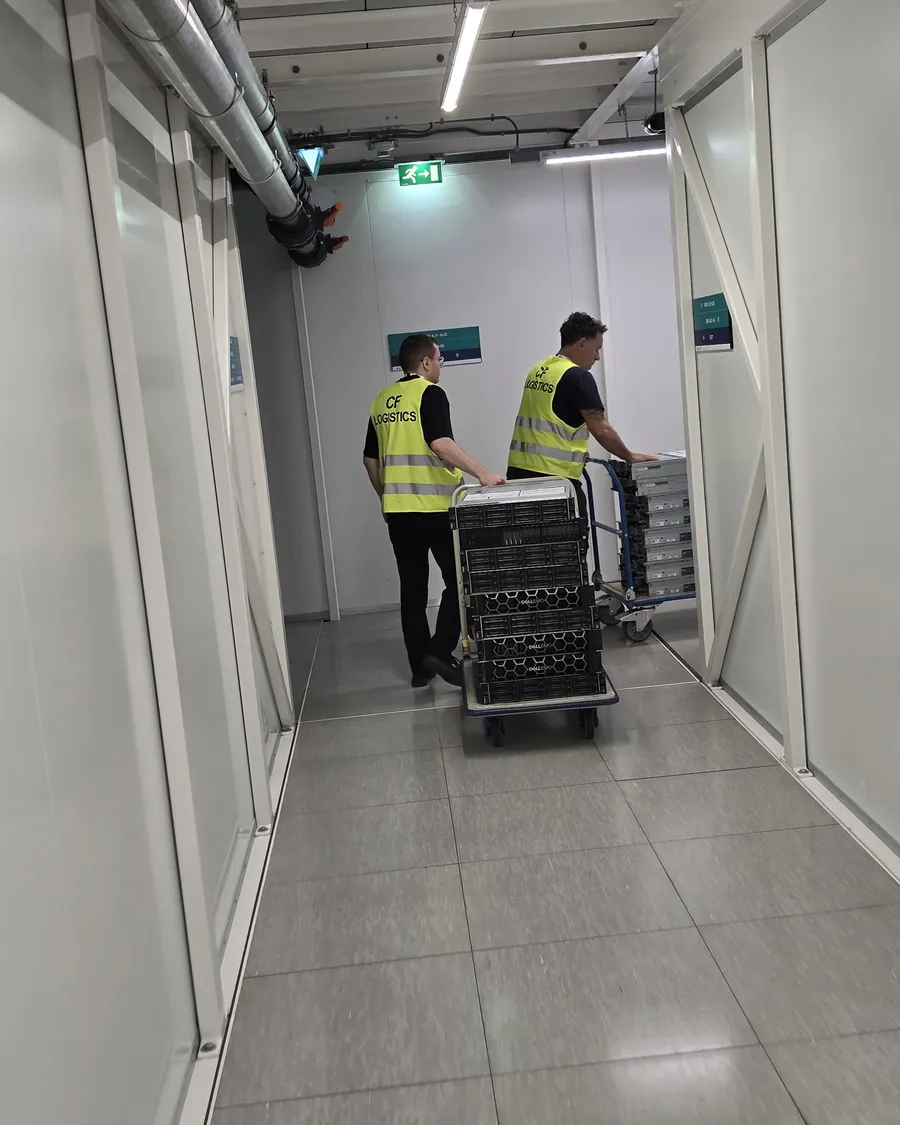 Two people in hi-vis vests pushing a trolley stacked with servers down a white data-centre corridor.