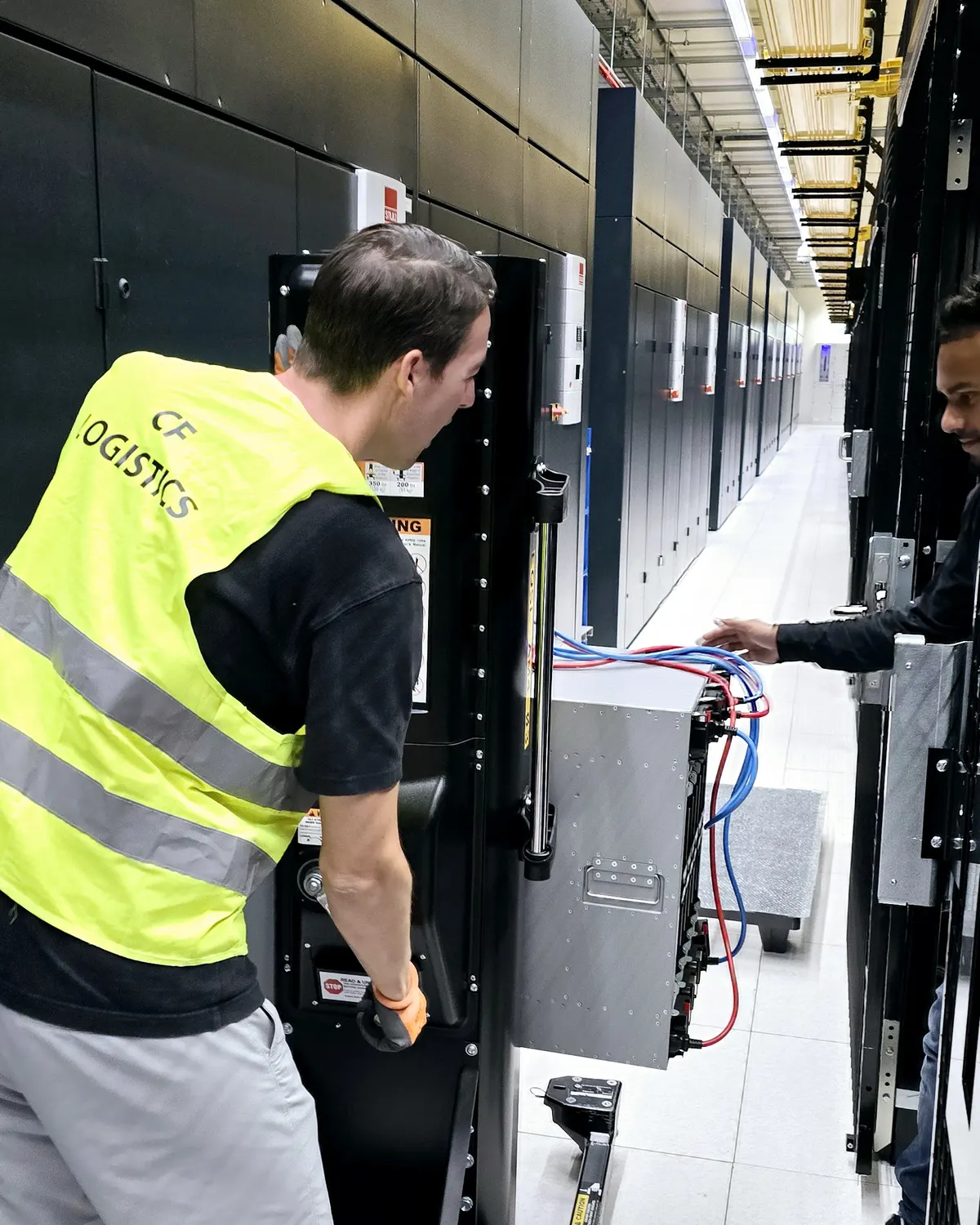 A worker in a CF Logistics hi-vis vest operating a server lift beside a rack in a data hall while a colleague works the cables.