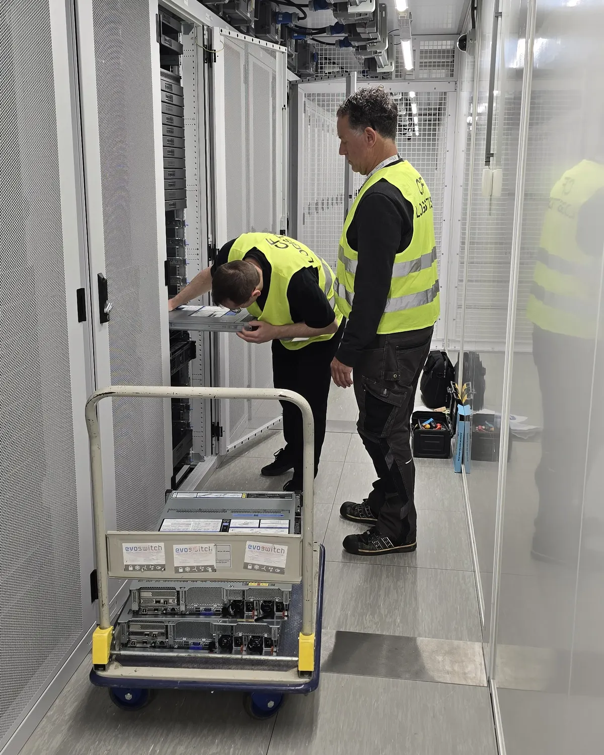 A worker in a hi-vis vest drawing a server out of an open rack while a colleague looks on, two servers already on a trolley beside them.