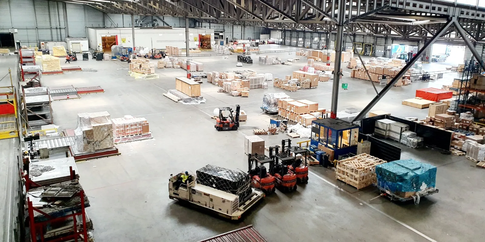 A freight hall seen from a mezzanine: crates and pallets across the floor, a row of forklifts, and a netted pallet on a dolly behind a tractor.
