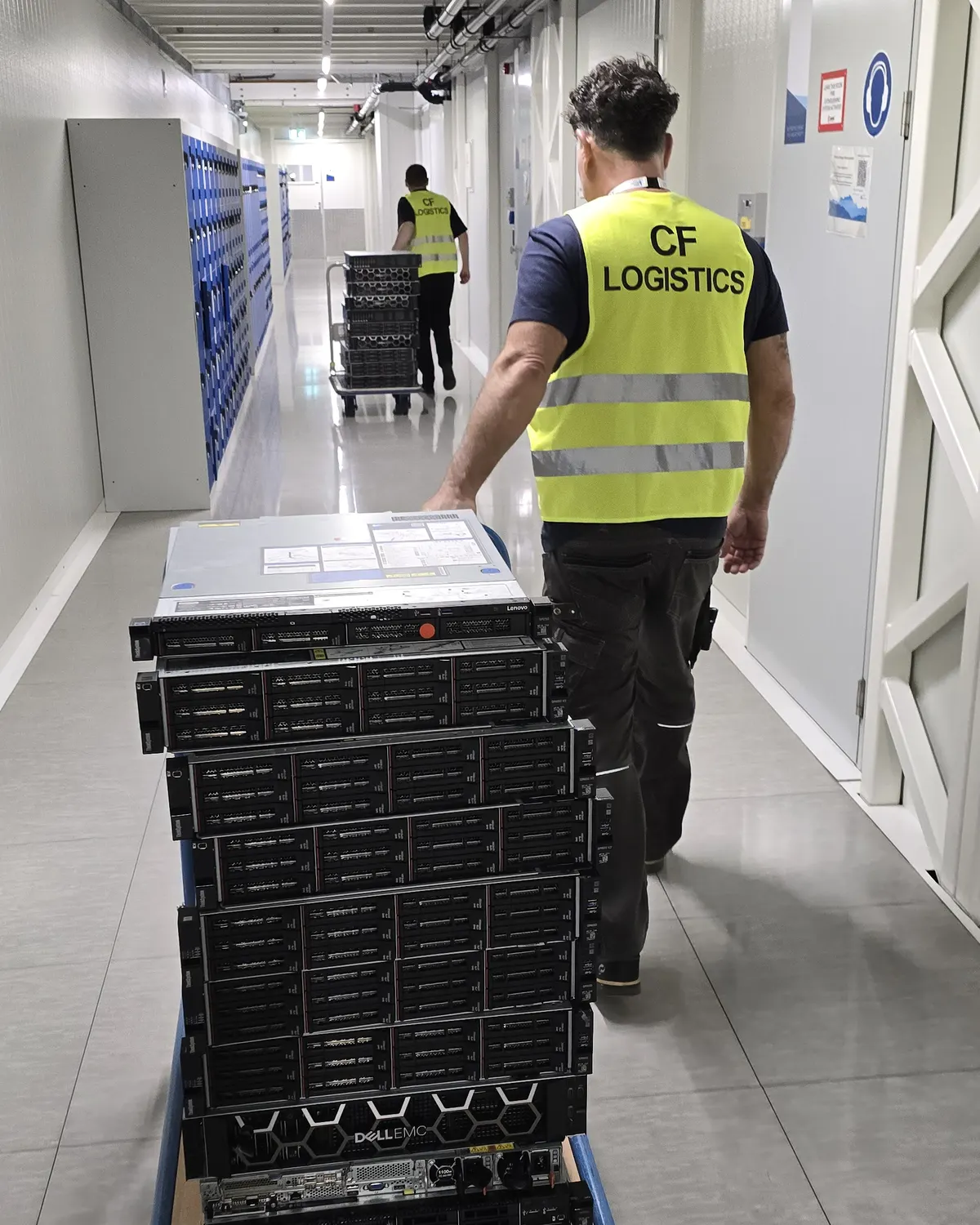A worker in a hi-vis vest pulling a trolley stacked with rack servers down a long white corridor, a second worker and trolley further ahead.