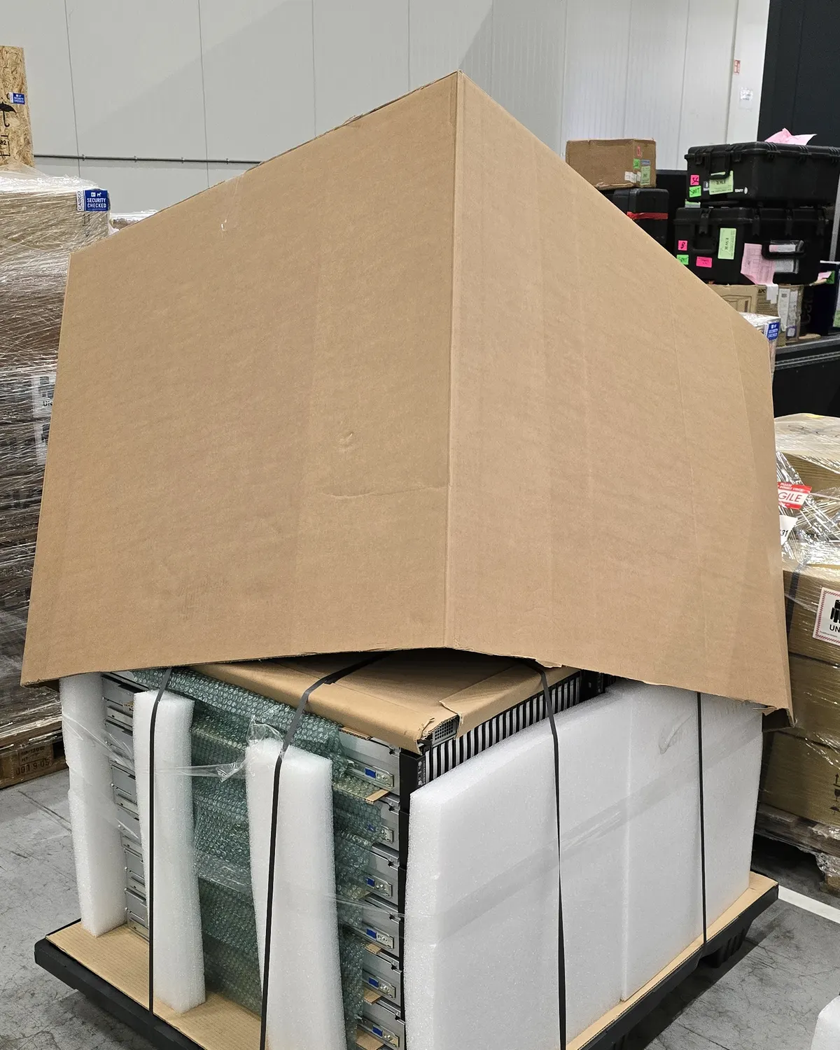 A cardboard carton being lowered over a strapped pallet of servers in bubble wrap and white foam corner pieces.