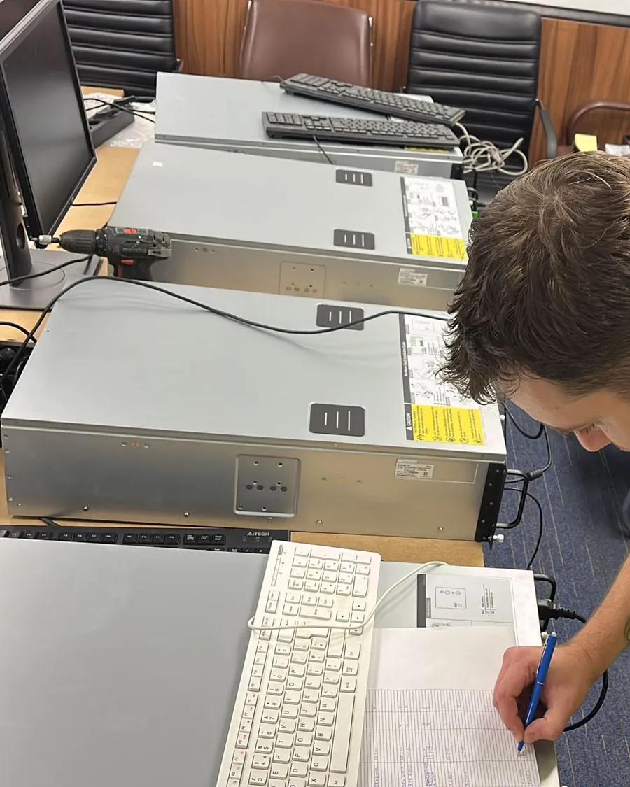 Servers laid out on a desk while a hand fills in a paper record beside them.