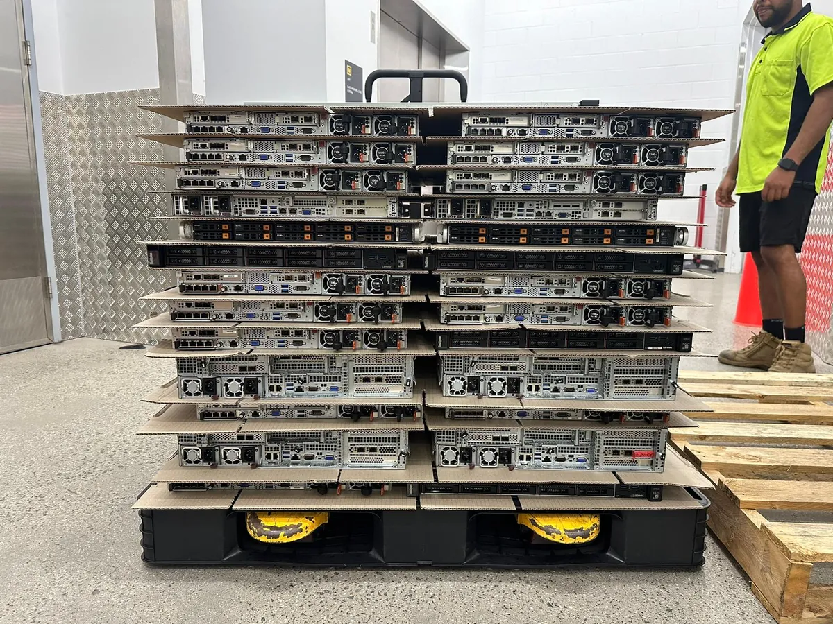 Rack servers stacked two wide on a black plastic pallet, a cardboard sheet between each layer, with a person in a hi-vis shirt standing beside them.
