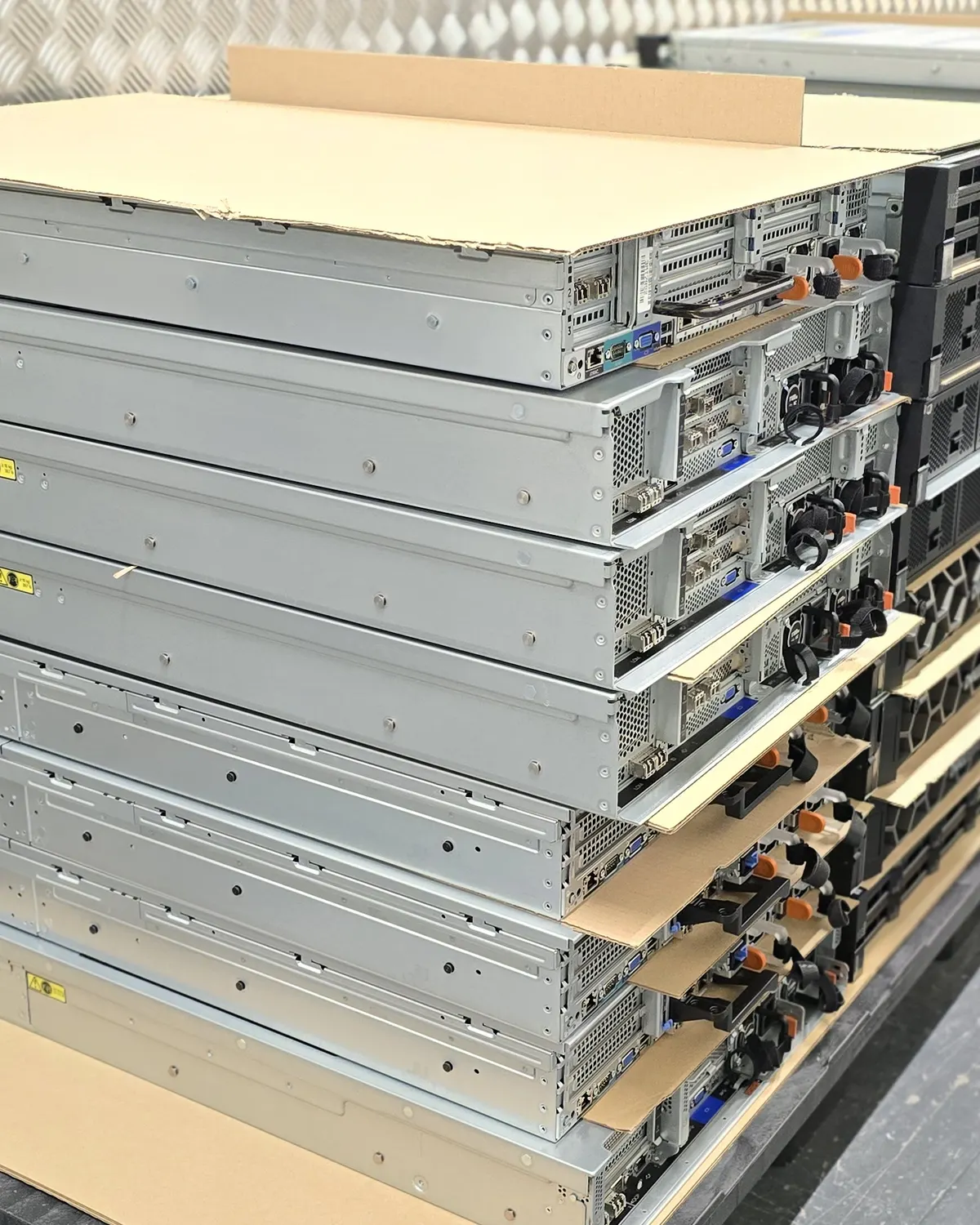 Rack servers stacked in a tall pile with sheets of cardboard between the layers, against a diamond-plate wall.