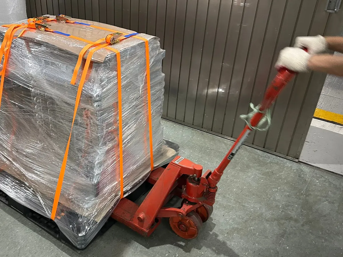 A gloved hand pulling a red hand pallet truck under a stretch-wrapped pallet held with orange straps.