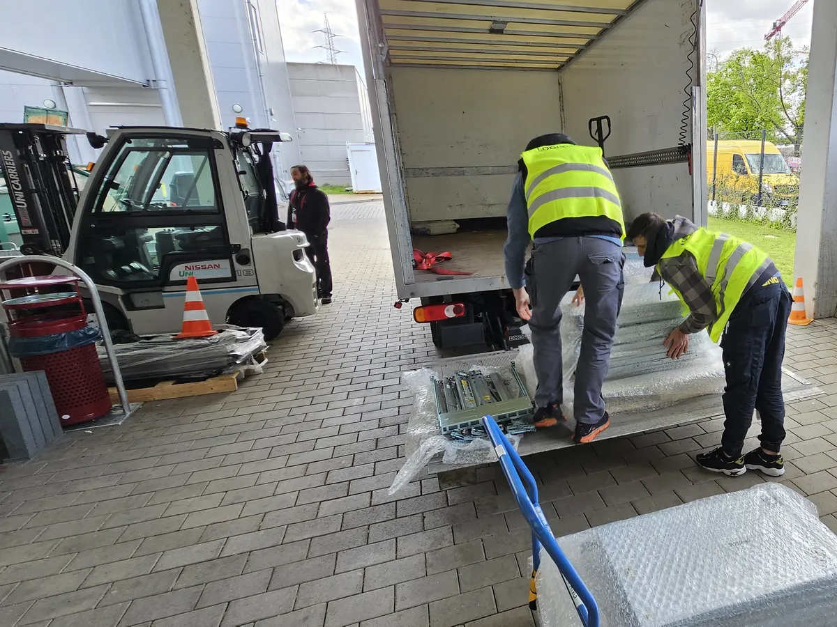 Two people in hi-vis vests unloading bubble-wrapped equipment from a box truck's tail lift onto a hand trolley.
