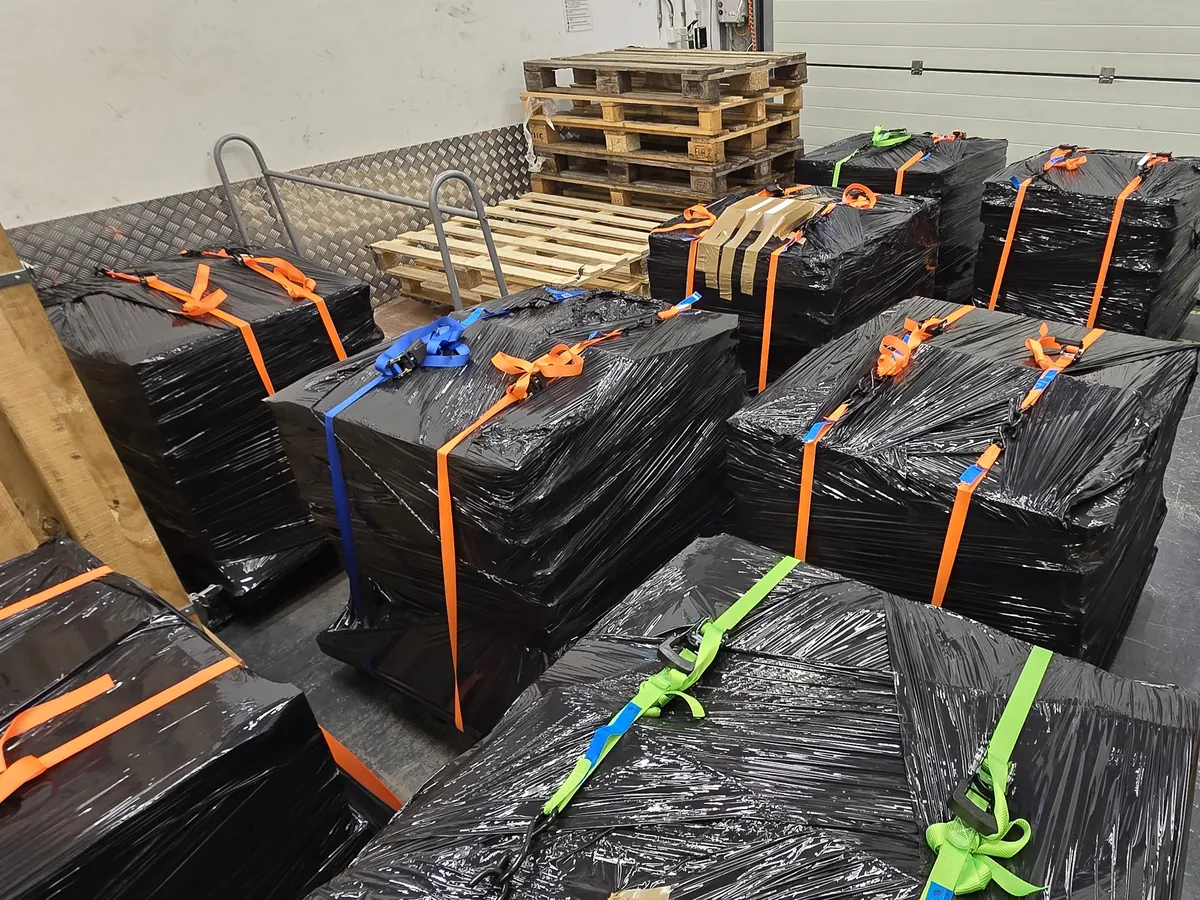 Black-wrapped pallets held down with orange, blue and green ratchet straps on a diamond-plate floor, empty pallets stacked behind.